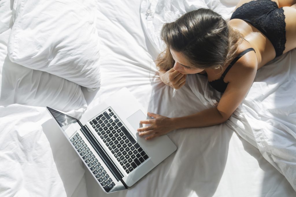 Frau, nur in Dessous begleitet, liegt auf dem Bett und schaut in den Labtop um sich im Netz mit Cyber-Sex zu vergnügen